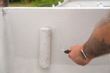 Close-up of a man hand holding a roller and painting a facade wall on a roof with gray paint. A handyman paints the facade of a house on the second floor in the summer against the backdrop of greenery