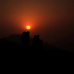 Fototapeta premium Individuals silhouetted against a warm yet slightly dark sunset while climbing a mountain.