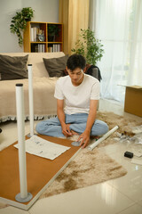 Young asian man sitting on soft rug assembling a table and following assembly instructions