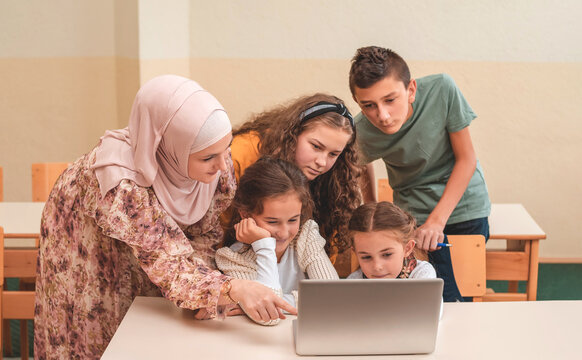 A female hijab teacher teaching group of child students using a laptop during the school class.
