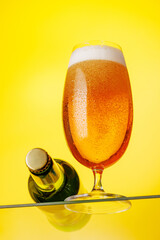 A tall glass of beer with a golden hue and frothy head, on a yellow background