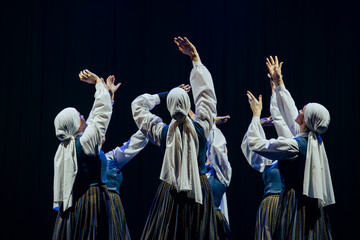 A group of dancers in traditional attire perform a synchronized and expressive routine on a dark...