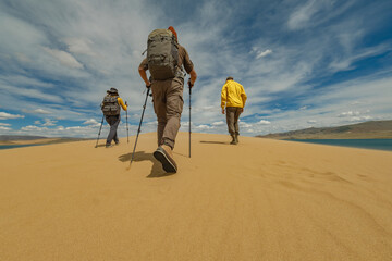 Travelers in bright clothes with backpacks on their backs walk along a sand dune against a backdrop of a lake and blue sky, showing off the soles of shoes in the sand.
