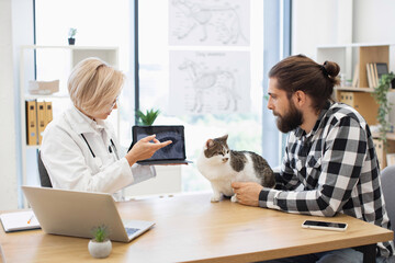 Veterinarian speaking to male pet owner while reviewing feline's x-ray. Scene shows caring...