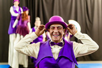 Cheerful circus performer in vibrant purple costume adjusts his hat, getting ready for a show, while other artists wait in the background