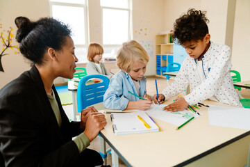 A female Black teacher sitting next to two multiethnic school-aged boys and looking while they are drawing together on a piece of paper, a White school-aged is sitting behind them in a classroom