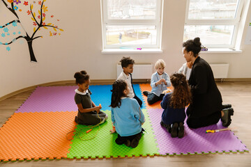 A female Latino teacher in her 30s with dark hair in a bun and diverse school-aged children,...