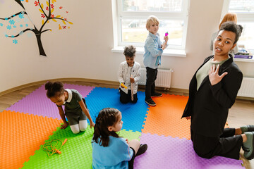 A Black female teacher in her late 20s, who is dressed in a black business suit, kneeling next to a...