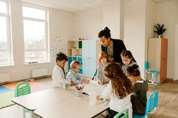 Obraz premium A Black female teacher stands at a table and talks to multiethnic primary school children who are sitting around it, drawing on a large sheet of paper, in a bright, spacious, and colorful classroom.