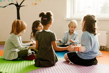 A diverse group of first-grade students, three girls, and three boys, sitting together in a circle on the mat in the corner of their classroom, holding hands and talking, as the sun shines on them