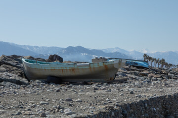 An old abandoned boat with rust stains rests on a rocky shore against a backdrop of snow-capped...