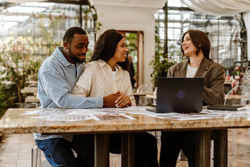 A happy couple of an adult Black man and a young Latino woman, engaging in conversation with an adult White female wedding assistant and using a laptop. They are sitting at a table in a spacious room.