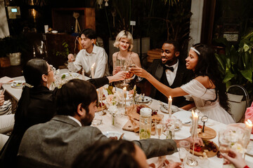 A group of diverse guests celebrates a wedding at a long dinner table with food. They toast with...