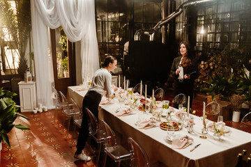 A European female wedding organizer in her 30s with brown hair with a clipboard standing in a wedding room next to a festive table and talking with a European adult female helpmate during preparation