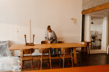 A young, overweight European woman in her late 20s, who is wearing a gray business suit, adjusting the chairs for a long wooden table, as she stands inside of a new, cozy coffee shop
