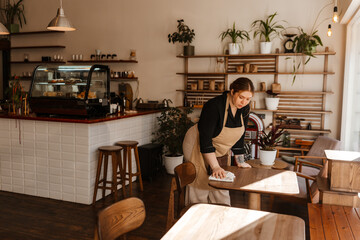 A White red-haired female employee in her late 20s, wearing a beige apron over a black shirt, is cleaning tables with a cloth, working in a sunlit, cozy coffee shop with a window to the right.