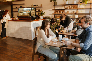 Diverse customers sit at tables, drinking and eating in a cozy cafe. An adult White female barista serves at the counter, with wooden shelves, plants, and a display case showcasing baked goods.