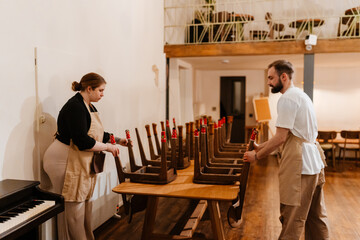 A White man in his early 30s and a young, overweight White woman in her late 20s, working together to place the chairs on top of the tables, as they're cleaning up in a small coffee shop after work