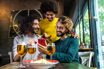 Three happy male friends using smartphone and drinking beer in restaurant