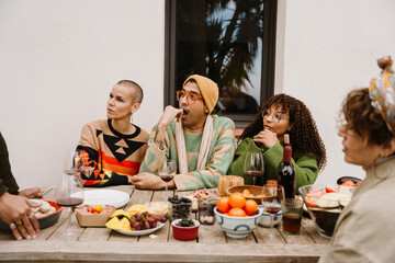 A group of diverse friends gathers for a meal, dishes, and drinks spread across the table as they enjoy each other's company. They are sitting in front of a white wall and looking to the left.