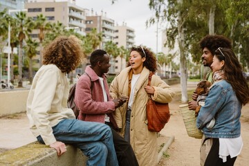 A group of diverse friends wearing casual clothes socializing in a city park, some standing, others sitting, engaged in lively conversation. An adult Latino woman talks to an adult Black man.