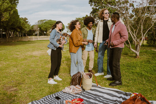 Happy diverse friends stand chatting in a grassy park with green trees. A picnic blanket with various items lies on the ground nearby. An adult tall White man with curly hair stands among his friends.