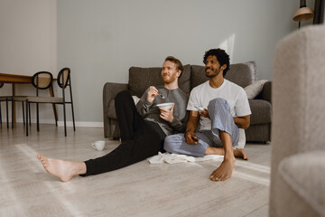 In a living room, a White brown-haired man and a Black brunet man, a gay couple in their 20s wearing home clothes, are sitting on the floor resting their backs on a sofa, eating, and watching TV.