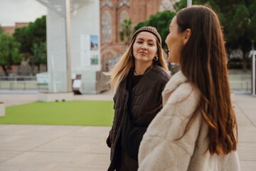 Outdoors, two adult White women walk and converse. One wears a hat and a puffer jacket, and the...