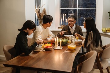 An elderly Asian grandpa in his late 70s, sitting at the head of a small table and having dinner with his family, his middle-aged daughter and her two adult grandsons, using chopsticks and chatting