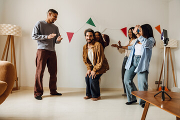 Five multiethnic friends, two men, and three women, are recording a funny video on a phone on a tripod on a table, dancing and laughing, in a living room decorated with colorful flags on the wall.