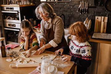 An elderly White grandma in her 60s, cutting shapes from dough with her two school-aged grandkids,...