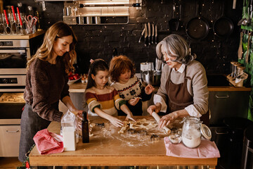 A White family consisting of an elderly grandmother, an adult daughter, and two school-aged grandchildren in sweaters standing around a table in the kitchen and cooking Christmas cookies together