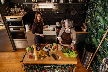 An elderly White mother in her late 60s cutting up some veggies and talking to her mid-30s White daughter, who helps her, as they stand together in a modern kitchen, with a black wall behind them