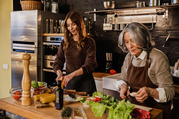 A Caucasian 30-year-old woman and her elderly mother dressed in aprons standing at a table in the kitchen with black walls, different colorful vegetables and dishes, and cooking together a meal