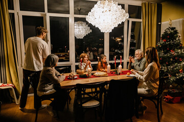 A White family of six, including the elderly grandparents and the school-aged kids, a boy, and a girl, sitting down in the evening to have Christmas dinner together, with a Christmas tree beside them