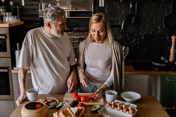 A European couple in their 50s stands at a wooden table in a kitchen, with the woman slicing tomatoes, surrounded by ingredients, engaging in a friendly conversation with the man on the left.