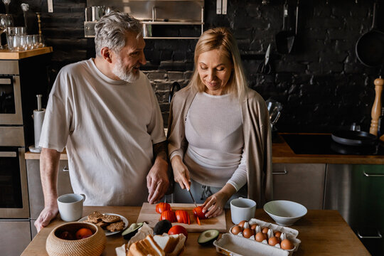 A European couple in their 50s stands at a wooden table in a kitchen, with the woman slicing tomatoes, surrounded by ingredients, engaging in a friendly conversation with the man on the left.