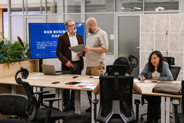 A multiethnic group of colleagues, two men stand and discuss documents, while two other women sit at the table during a business meeting, with a big monitor and lockers behind them, in an office.