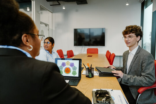 A group of multiethnic colleagues participates in a meeting in a modern office, with a laptop displaying "7 Steps to Boost Social Media Engagement" as they discuss work.