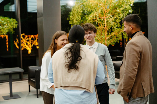 A young White man in his 20s, talking to a diverse group of his three young colleagues as they stand together in a small circle inside of an office hall, with small potted trees in the background