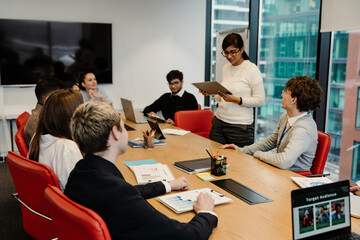 A group of diverse colleagues collaborates in an office room with big windows, reviewing documents and using digital devices. A young South Asian woman in glasses stands and gives a presentation.