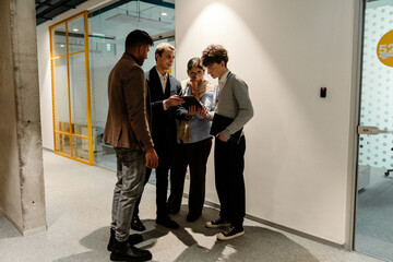 A White businessman in his 30s, who is dressed in a black business suit, showing his tablet to a diverse group of young interns, as they stand together in an office hallway