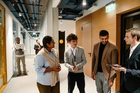 In a modern office hall, a multiethnic group of formally dressed colleagues are talking to each other, one of them is a young intern, a White man in his early 20s wearing a gray cardigan and glasses.