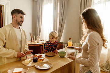 In a bright kitchen, a smiling White family prepares their breakfast at a counter, with the man in his 30s preparing sandwiches, next to his school-aged son, and his wife in her 30s holding a jar.