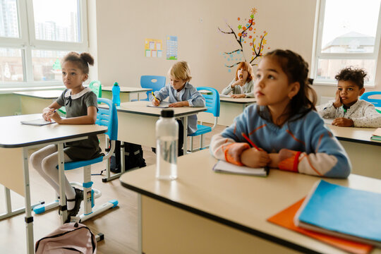 A young, school-aged White boy who is dressed in a button-up shirt, sitting at his desk in a first-grade classroom, writing in his notebook, as his diverse classmates look forward