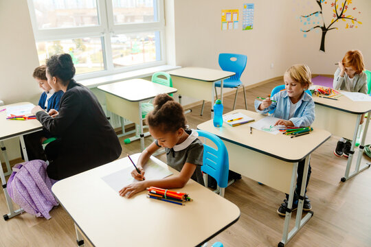 A young, school-aged Black girl, sitting at her desk in a classroom and drawing something on her paper, as her mid-30s Black female teacher helps one of the other students, in school