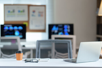 Modern office desk with laptop, coffee, eyeglasses and notebook, suggesting a productive and successful work environment, with blurred background showing charts and graphs on computer screens