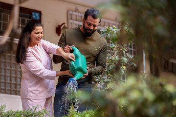 Young indian couple watering plants together outside their house garden in summer.Husband wife...