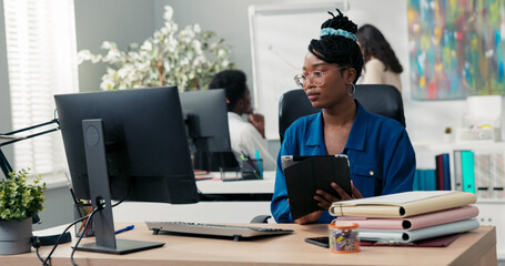 A professional middle-aged woman sits at her desk in a corporate office, holding a tablet,...
