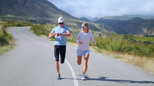 Majestic mountains with green slopes and a clear blue sky form an inspiring backdrop as two professional athletes run along a winding road. The man checks his smartwatch, tracking the distance.